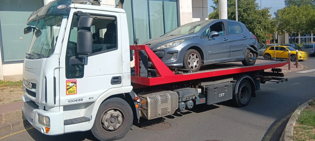 Porte-voiture blanc et rouge chargé d'une Peugeot 308 grise tenue par des sangles, bureaux et logements de Créteil derrière